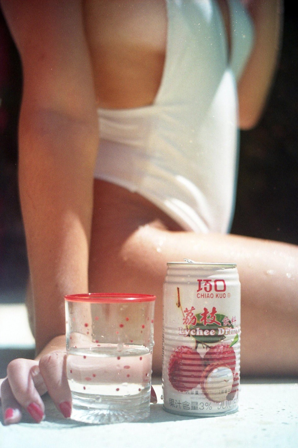 A woman in a white swimsuit holds a glass of water next to a can of lychee drink.