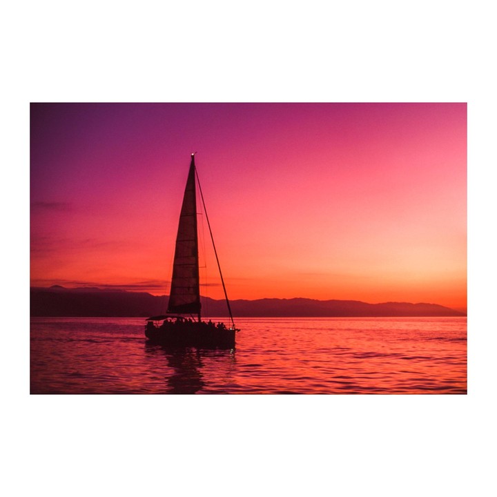 A sailboat with people on board is silhouetted against a vibrant pink and orange sunset over water with mountains in the distance.