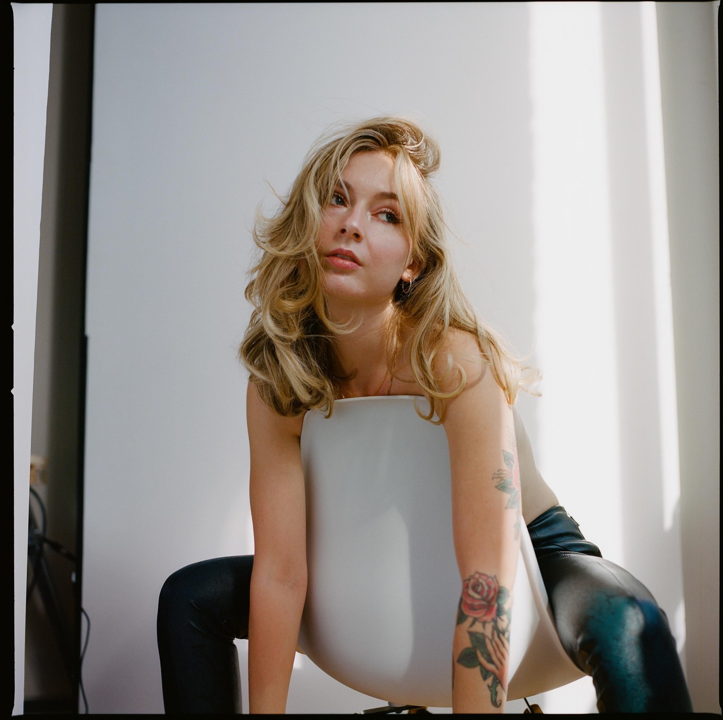 A woman with long blonde hair and a tattoo on her arm sits backward on a white chair.