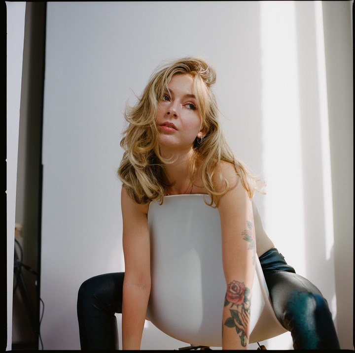 A woman with long blonde hair and a tattoo on her arm sits backward on a white chair.