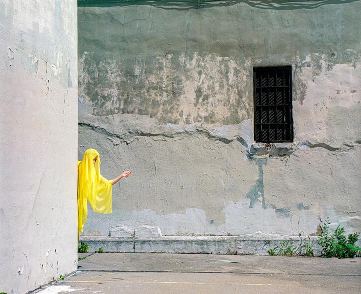 A person in a yellow veil and robe extends an arm from behind a wall towards a window with bars.