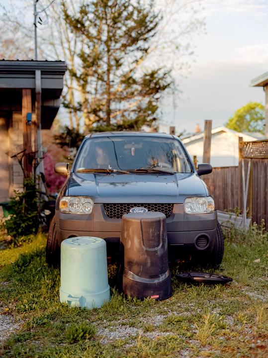 A dark gray Ford SUV is parked on a gravel and grass patch with two overturned bins in front of it.