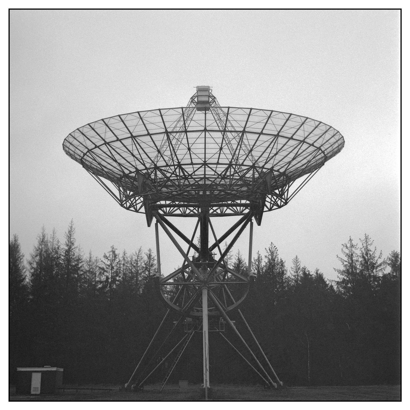 A large radio telescope stands above a line of trees under a cloudy sky.