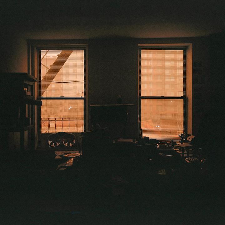 A dark room is illuminated by orange light filtering through two windows with blinds.