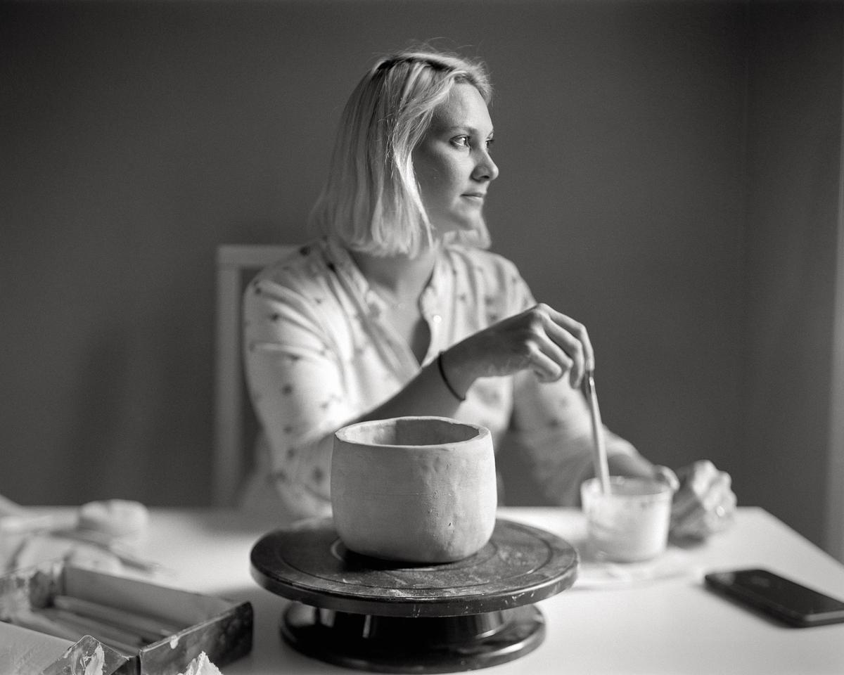 A woman sits at a table working on a clay pot.