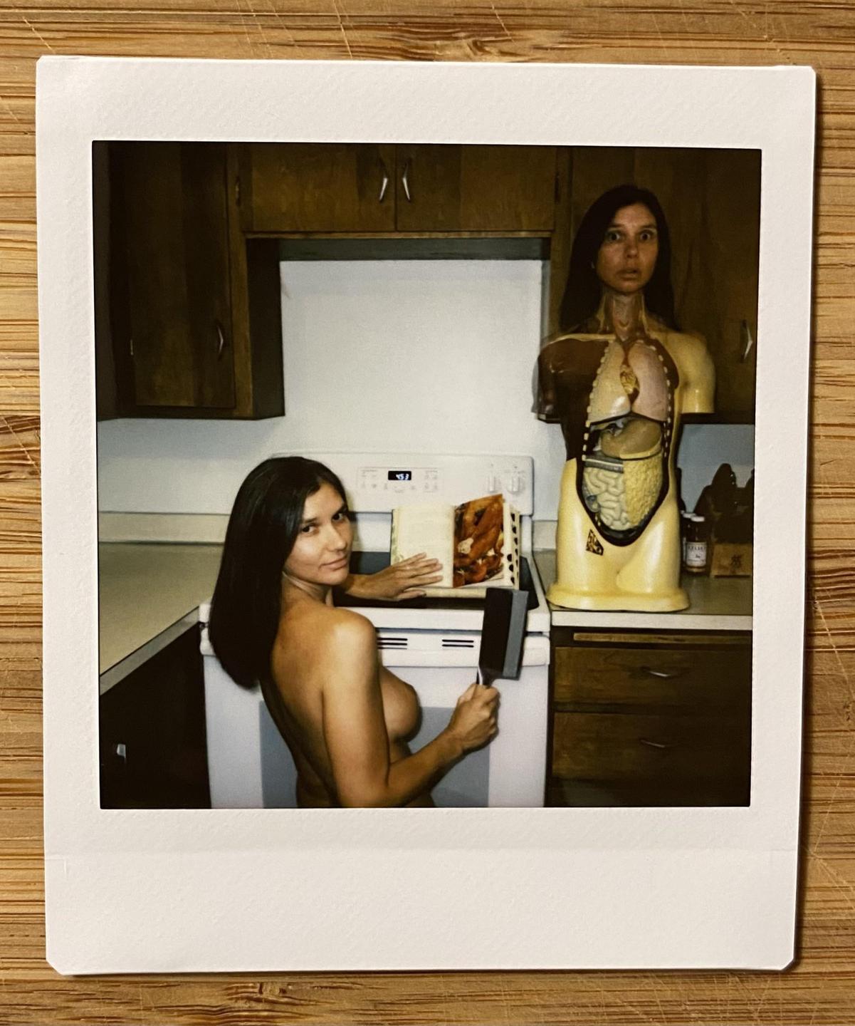 A topless woman holds a spatula in a kitchen next to an anatomical model.