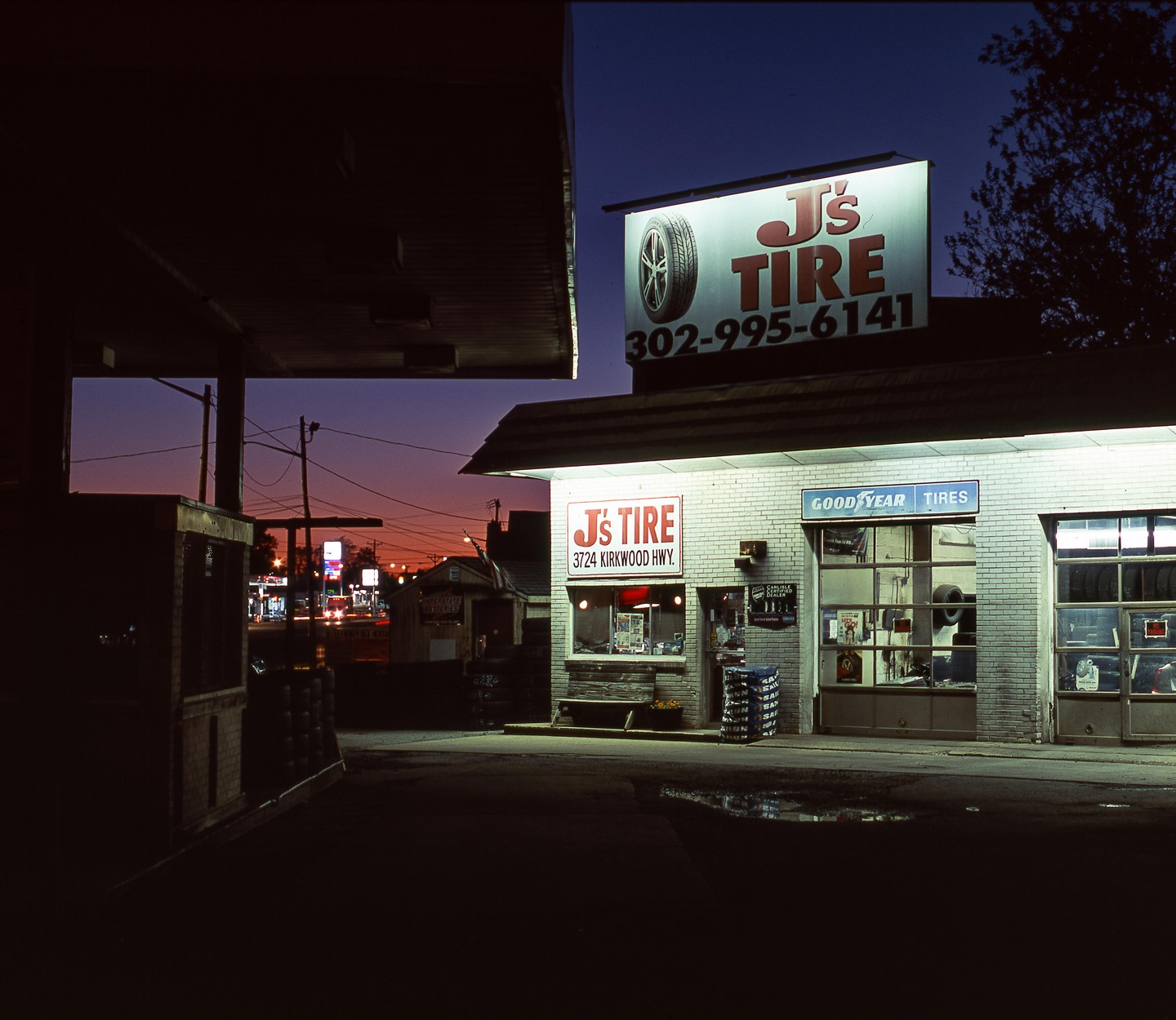 A tire shop with illuminated signs stands under a dark sky with a colorful horizon.