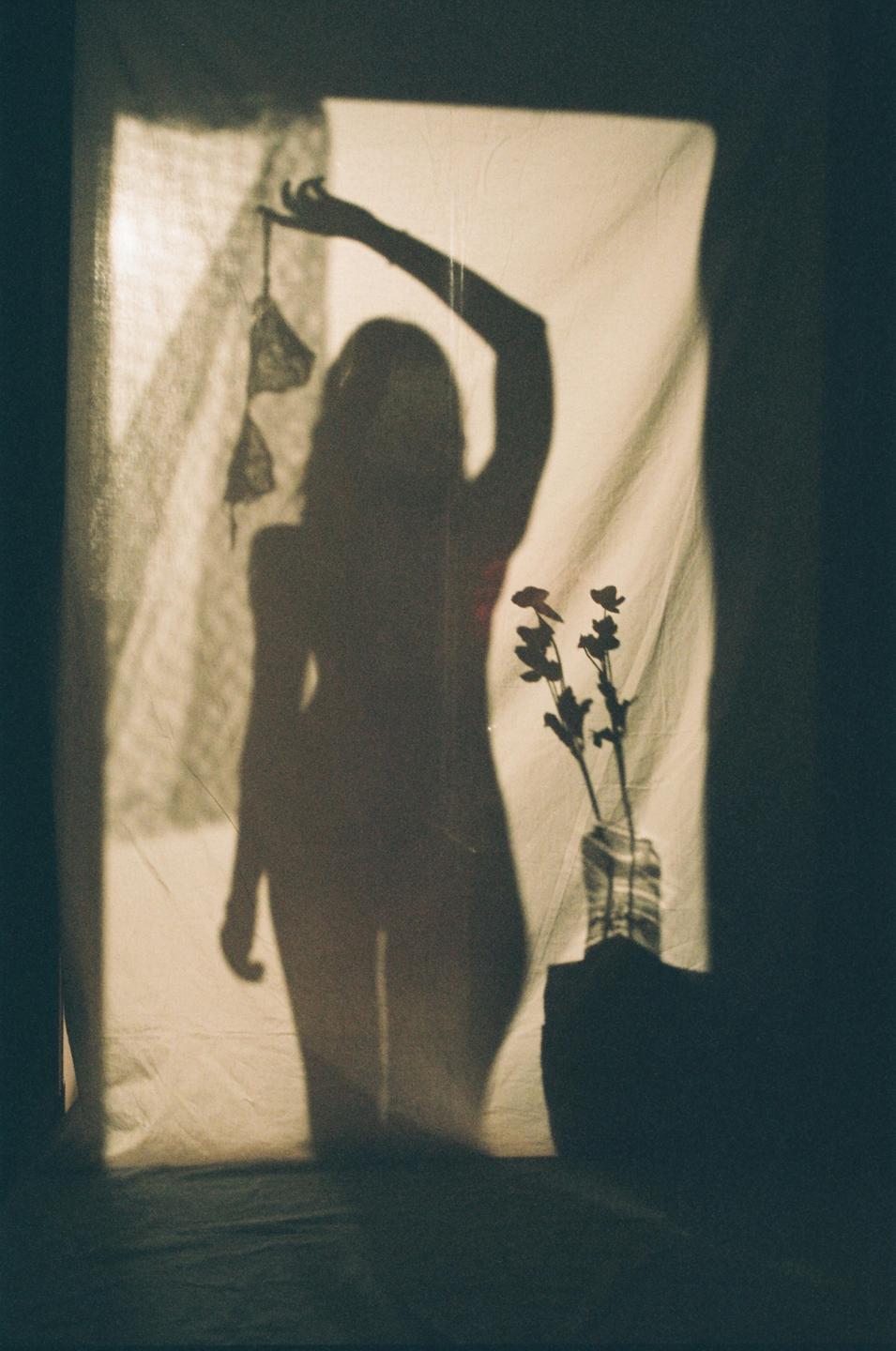 A woman's shadow is cast on a sheet, holding up a bra, with a vase of flowers to her right.