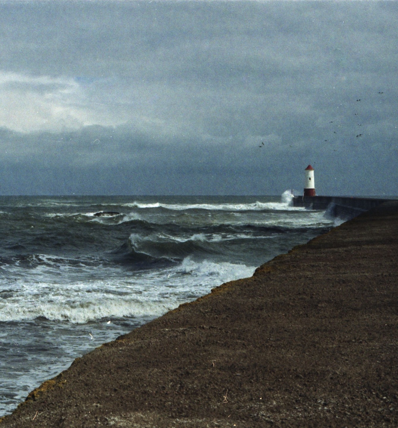 A lighthouse stands on a pier as waves crash against it under a stormy sky.