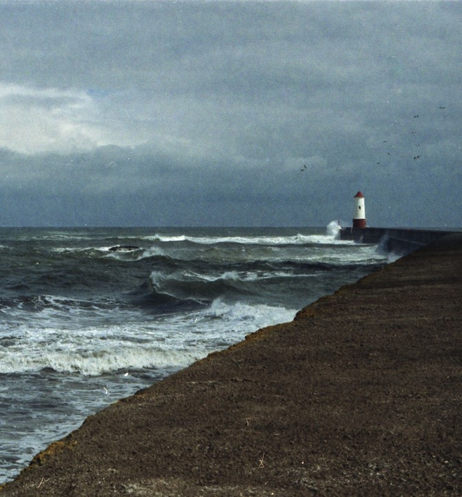 A lighthouse stands on a pier as waves crash against it under a stormy sky.