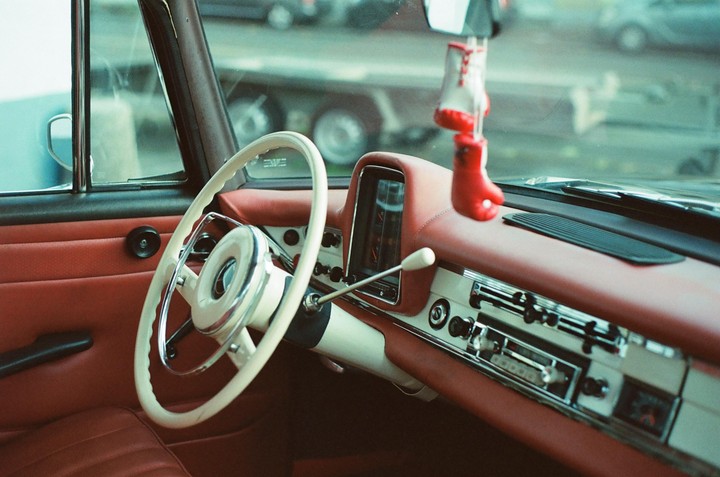 The interior of a vintage car features a white steering wheel, red dashboard, and miniature boxing gloves hanging from the rearview mirror.