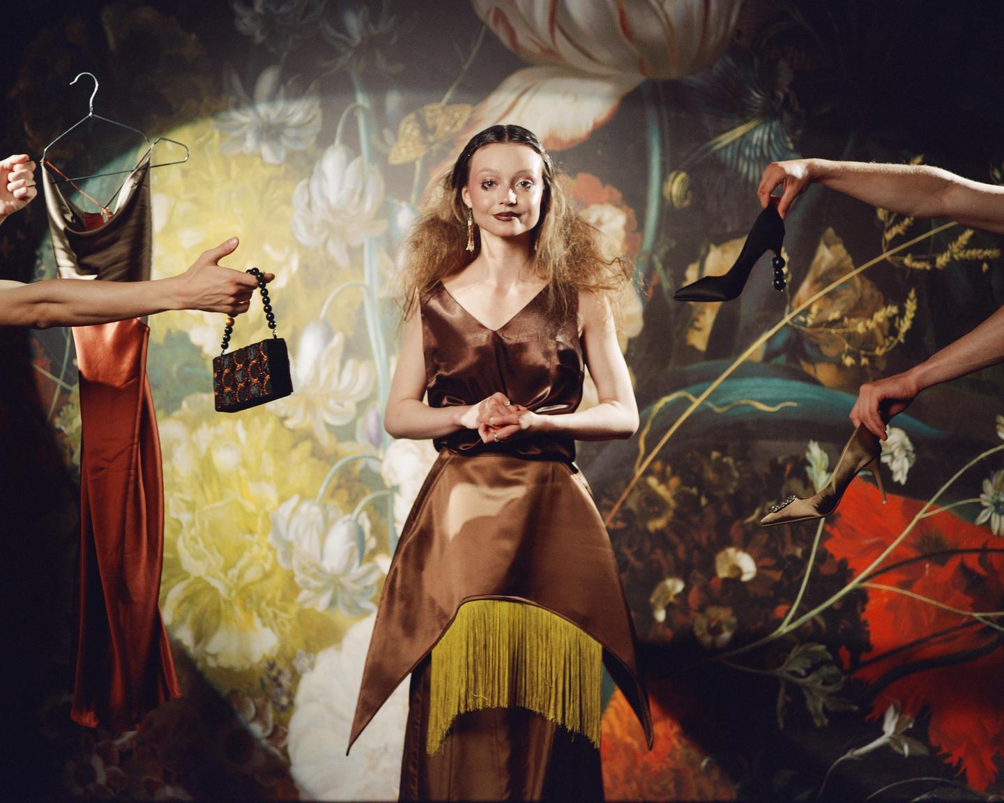 A woman in a brown dress with yellow fringe stands in front of a floral backdrop while disembodied hands offer her a dress, a handbag, and two high heels.