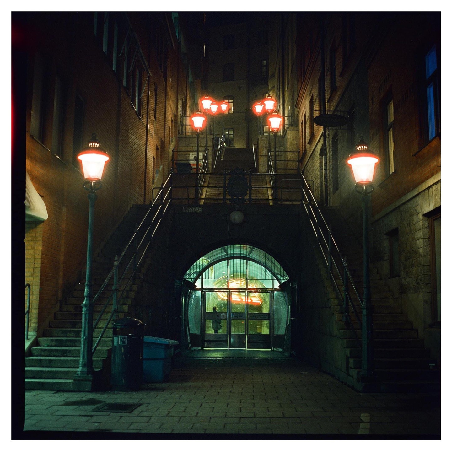 A narrow street at night is illuminated by red streetlights and a glowing tunnel entrance.