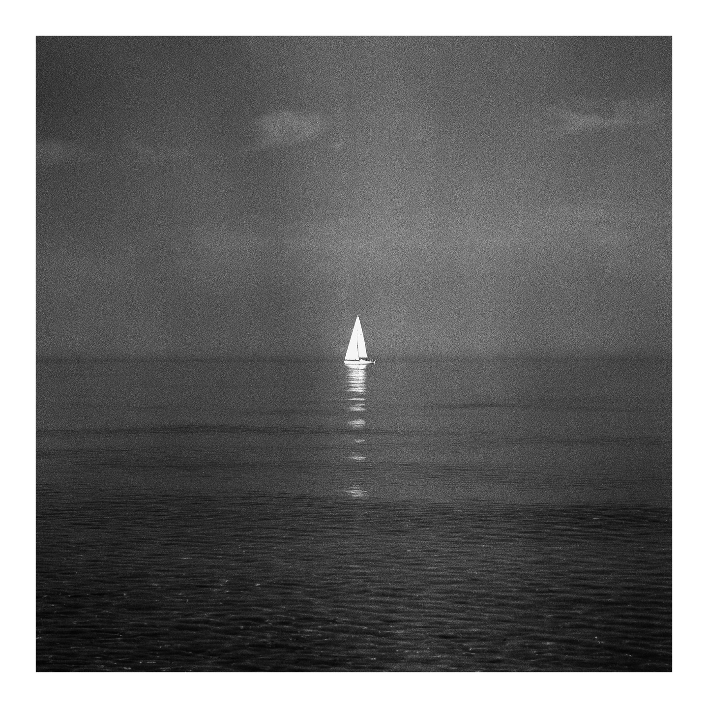 A sailboat with a bright white sail floats on calm water under a dark sky.