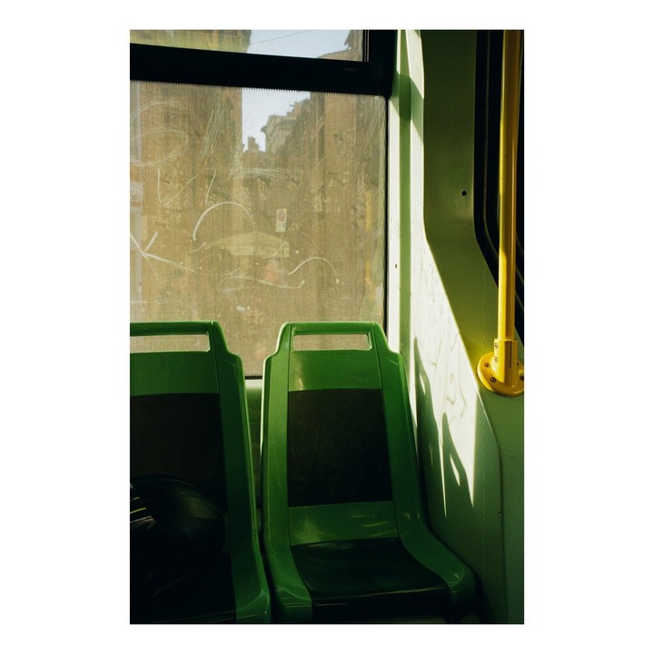 Sunlight streams through a dirty window onto two green seats and a yellow pole inside a vehicle.
