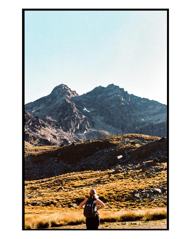 A woman with a backpack stands in a field of golden grass, looking towards a mountain range under a clear sky.