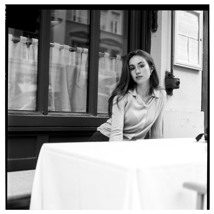 A woman sits at a table outside a building with a window and a menu.