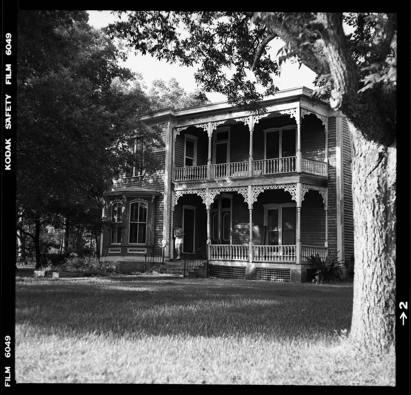 A large, ornate house with two stories of porches and balconies stands behind a grassy lawn, partially obscured by trees.