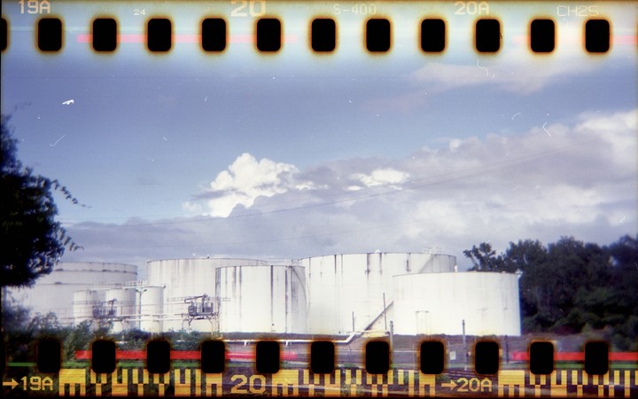 Several large white industrial tanks are visible behind a line of trees under a cloudy sky.