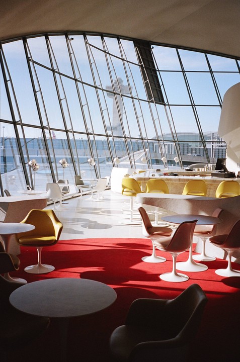 A lounge area with modern furniture and a red carpet is visible through a large glass window overlooking an airport control tower.