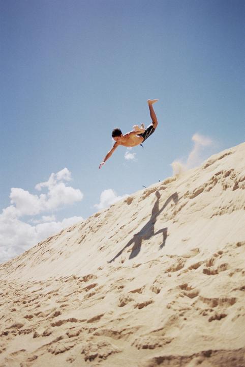 A man jumps in the air above a sand dune, casting a shadow on the sand.