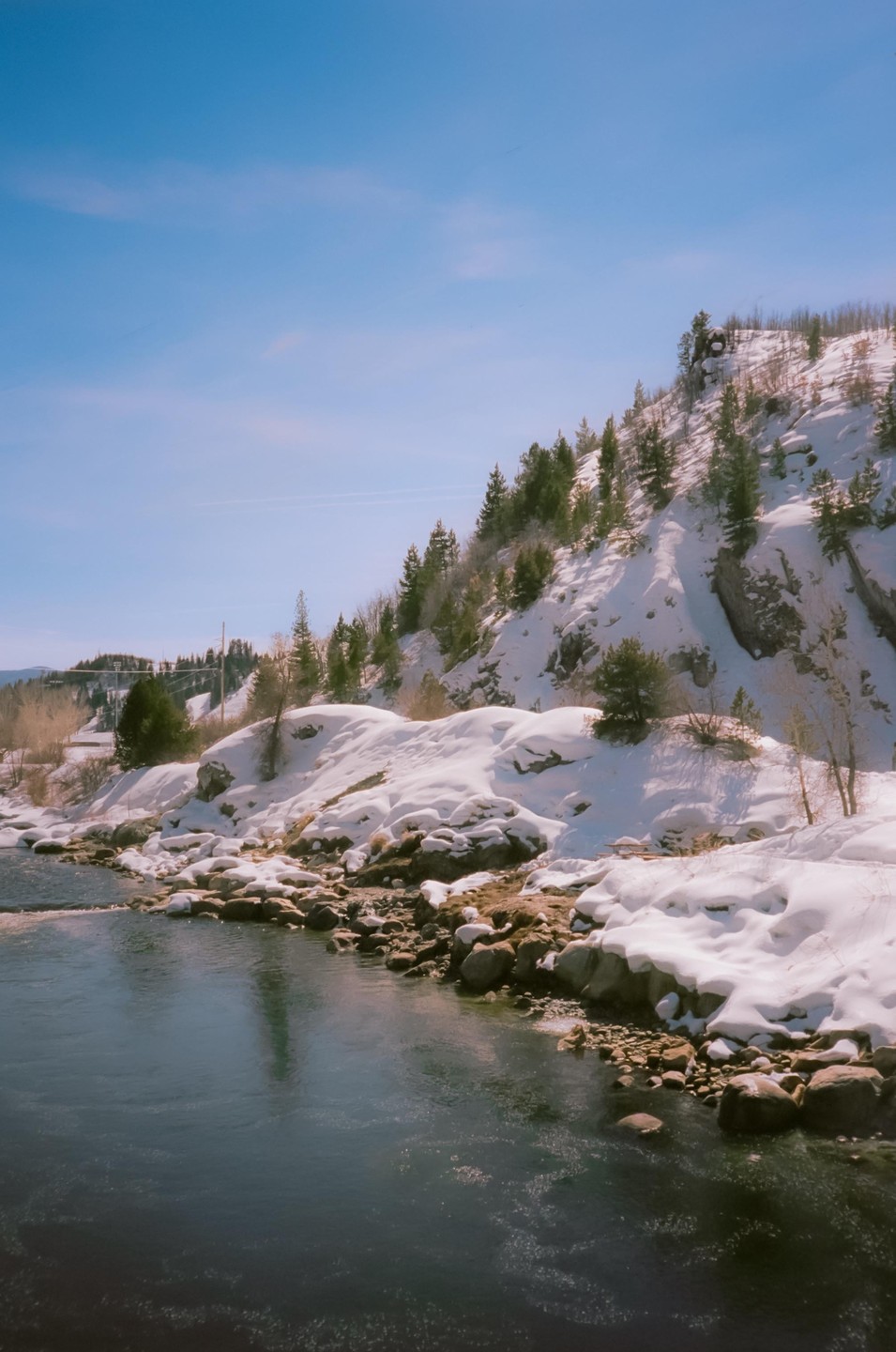 A river flows past a snow-covered bank with evergreen trees on a hill under a clear sky.