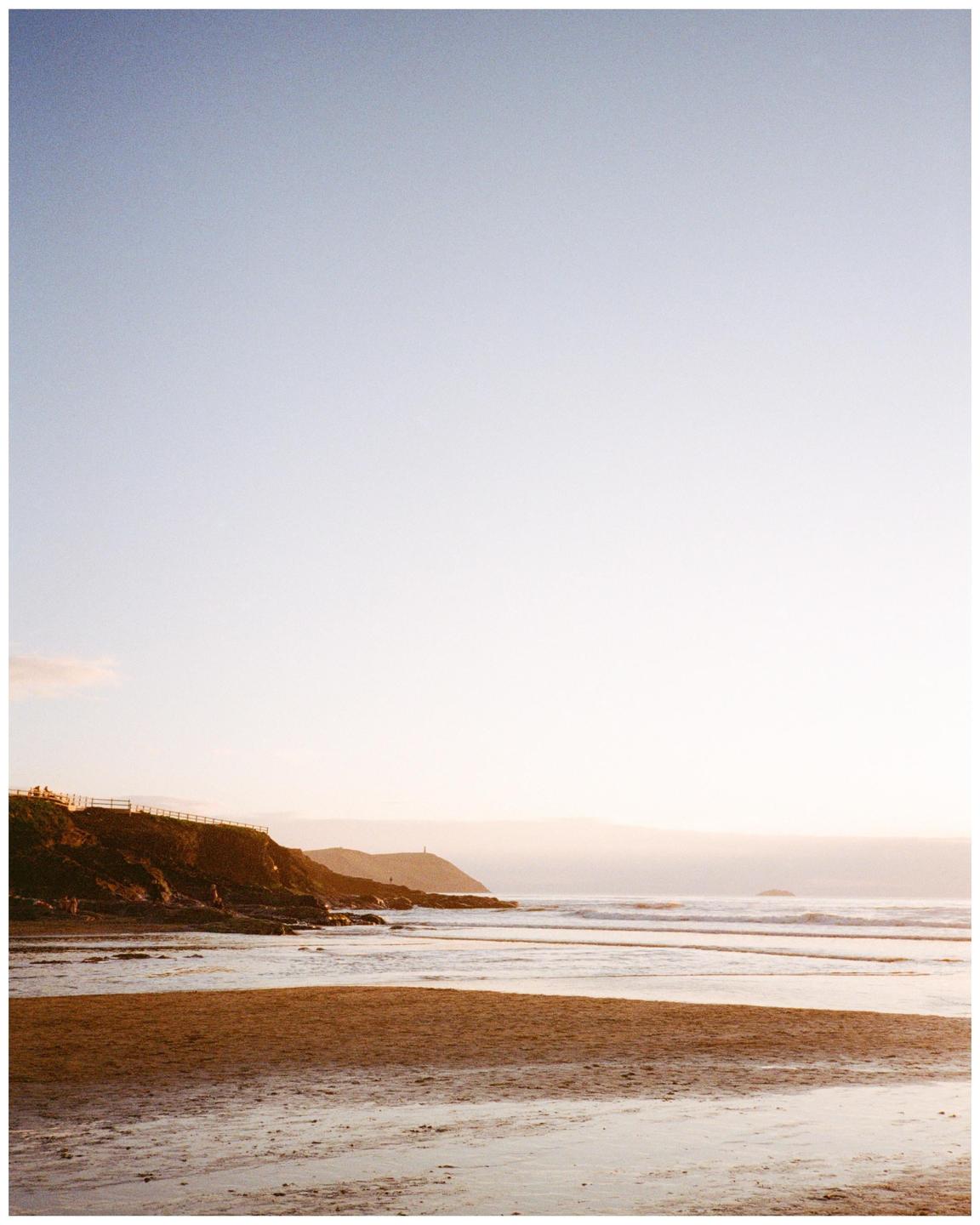 A sandy beach with waves crashing on the shore next to a rocky cliff under a clear sky.