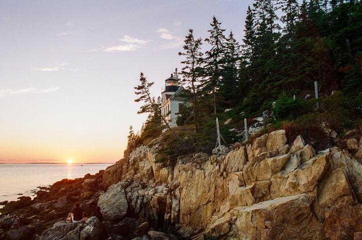 A lighthouse stands on a rocky cliff overlooking the ocean at sunset, with people sitting on the rocks in the foreground.
