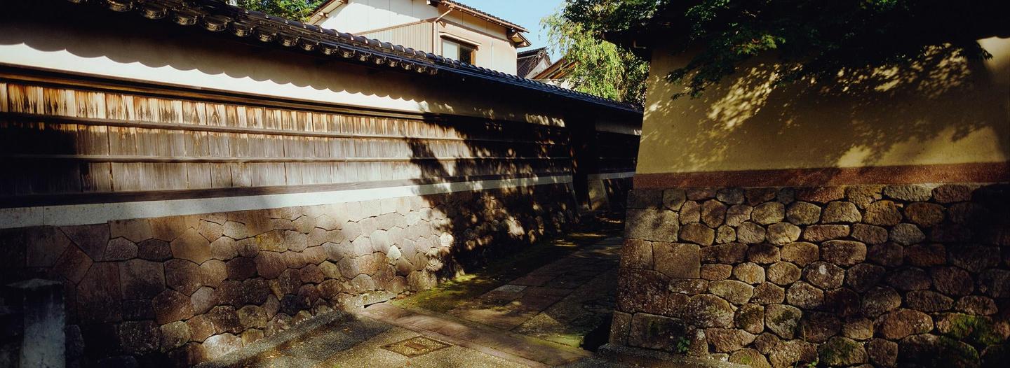 Sunlight casts shadows of leaves onto a stone and wooden wall along a narrow path in Japan.