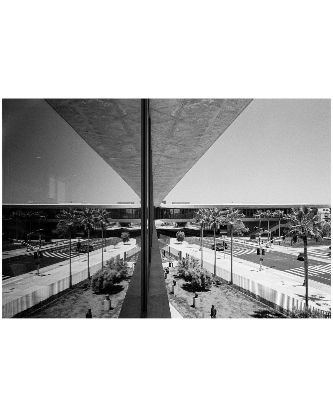 A black and white photograph shows a building with a large overhang reflected in a glass facade, with palm trees and a street visible below.
