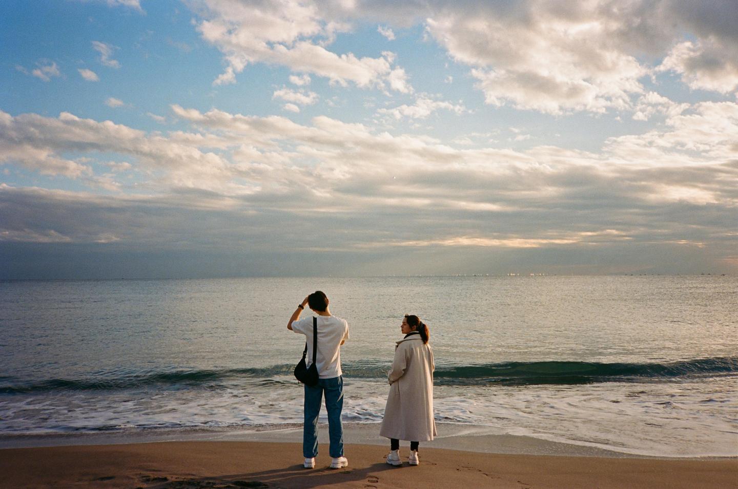 A couple stands on a sandy beach looking out at the ocean under a cloudy sky.