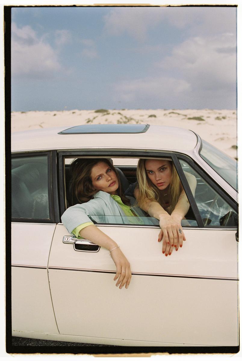 Two women are looking out of the windows of a white car in a desert landscape.