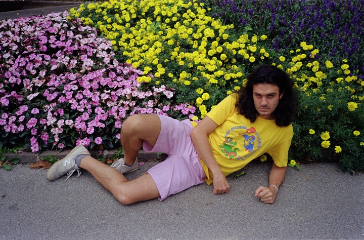 A man with long, dark hair lies on the ground next to a flower bed with pink, yellow, and purple flowers.