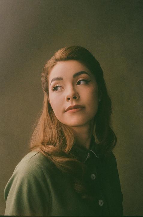A woman with long hair and a green shirt looks to the side against a green background.