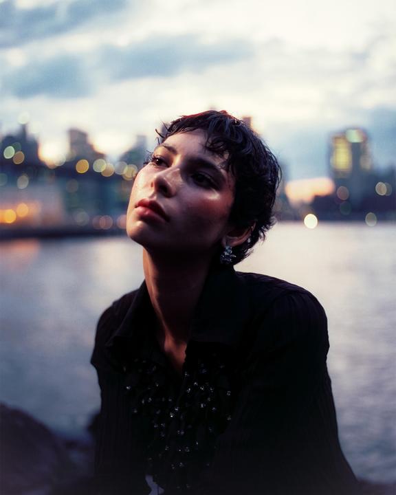 A woman with short, dark hair looks up with a city skyline and water in the background at dusk.