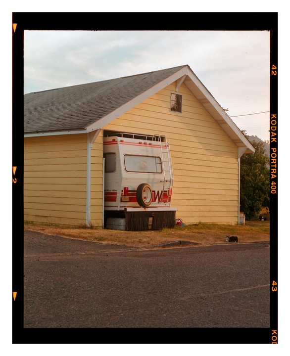 A white camper is parked under a yellow house with a cat sitting on the grass nearby.