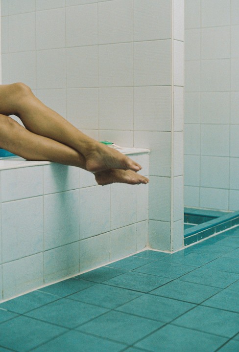 A person's legs and feet are visible above a tiled floor and bathtub in a bathroom.