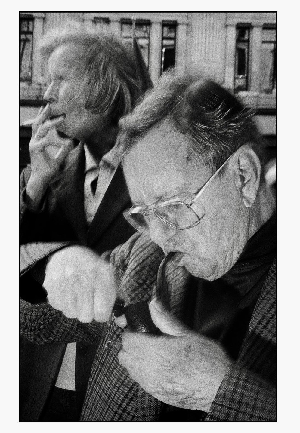 A man with glasses lights a pipe while a woman smokes a cigarette in the background.
