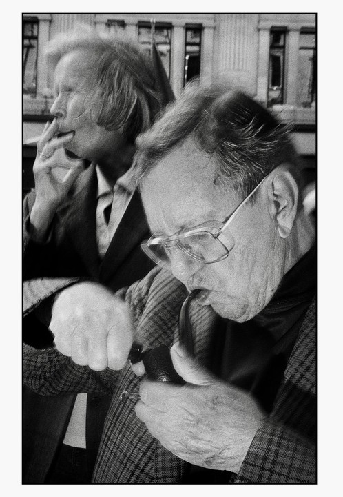 A man with glasses lights a pipe while a woman smokes a cigarette in the background.