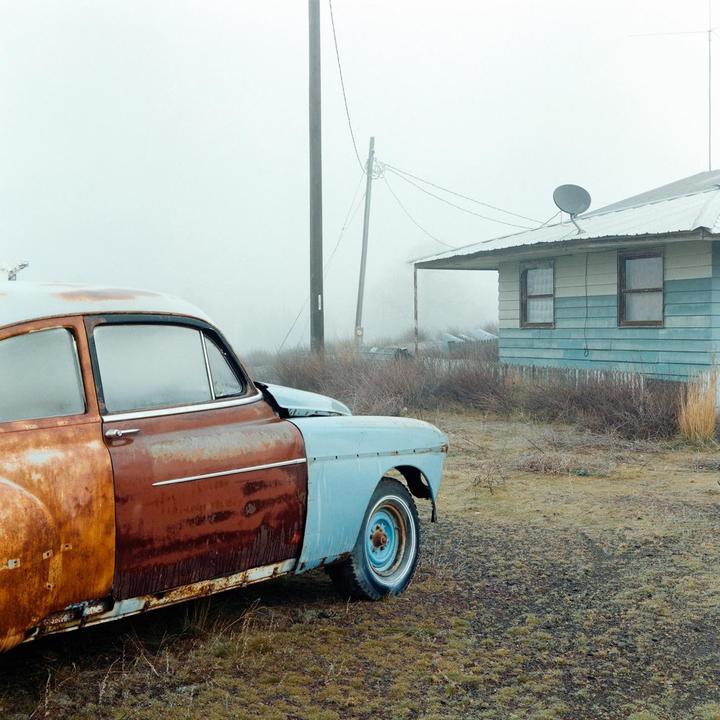 A rusty old car is parked in front of a house on a foggy morning.
