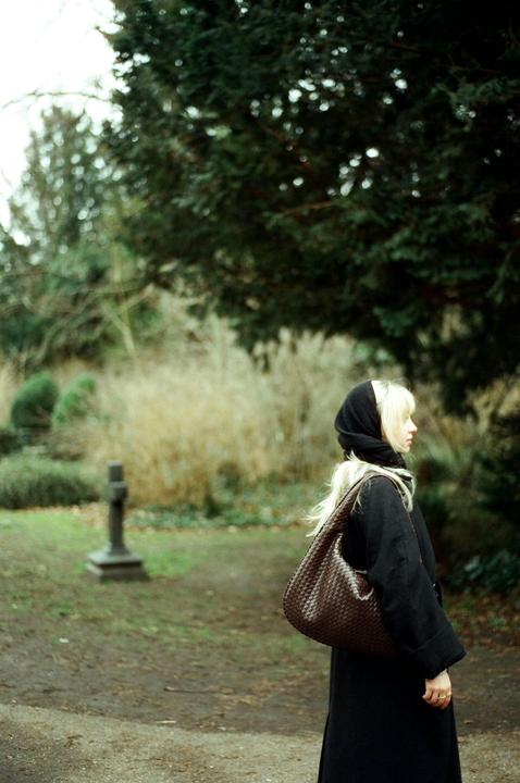 A woman with blonde hair and a black headscarf stands in a park with a stone cross in the background.