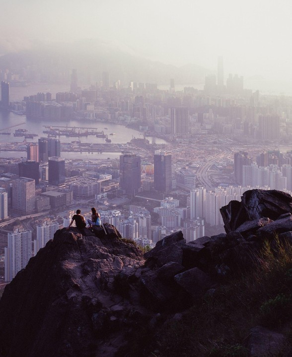 A couple sits on a rocky outcrop overlooking a hazy city with a body of water.