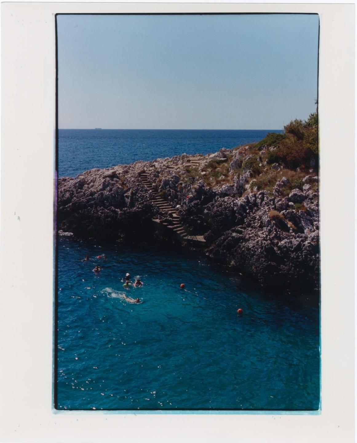 A group of people swim in a clear blue cove next to a rocky shore with stairs leading up a hill.
