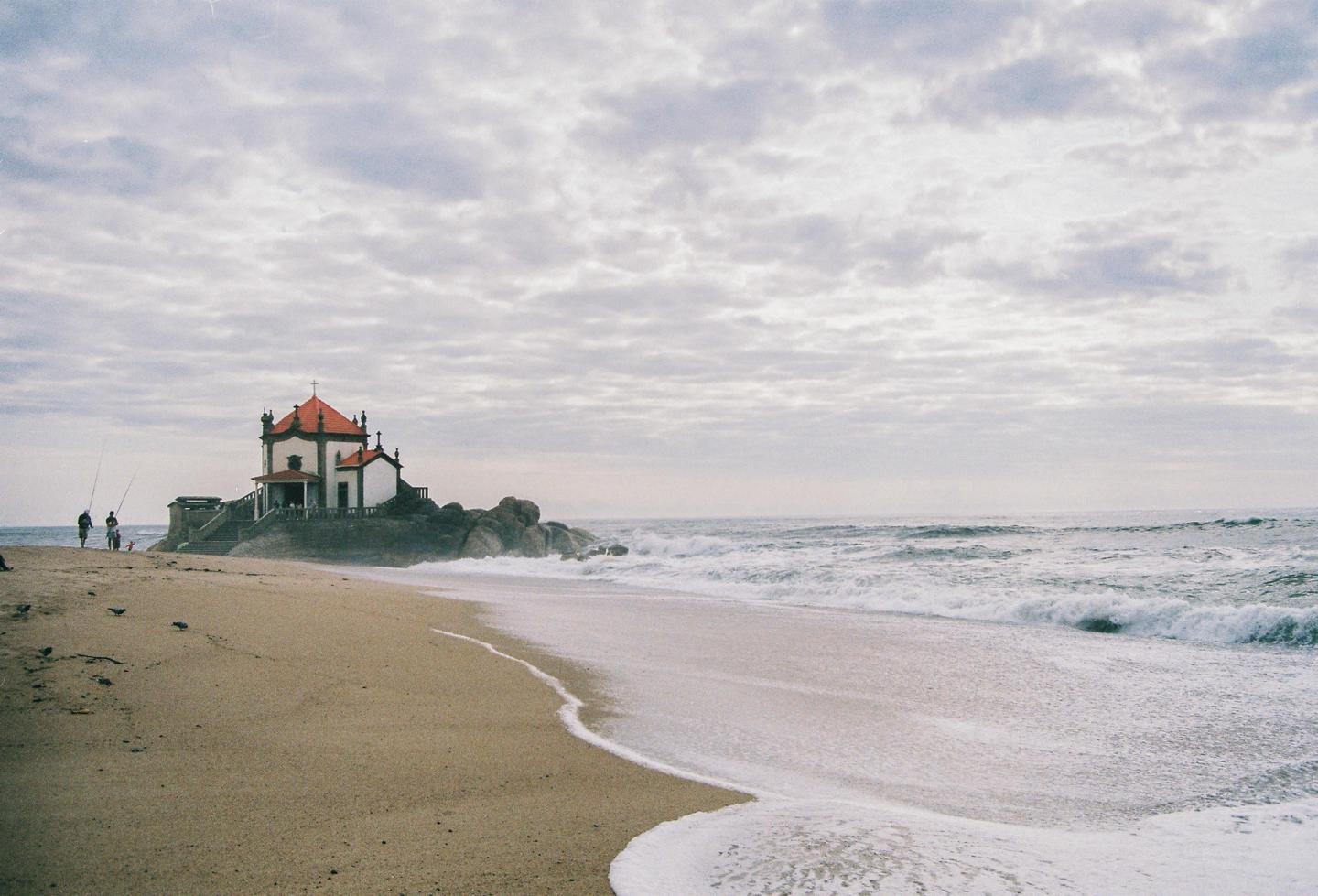 A small church with a red roof sits on a rocky outcrop next to a sandy beach with waves crashing on the shore.