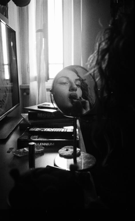 A woman applies lipstick while looking into a round mirror on a cluttered table next to a window.
