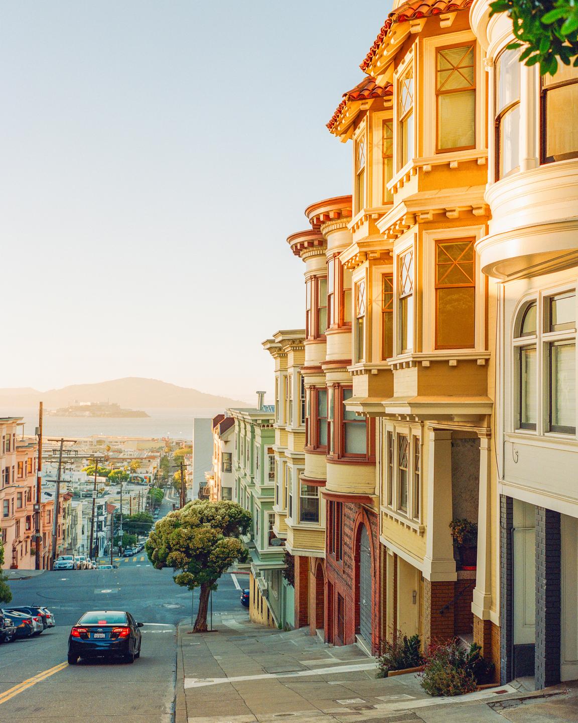 A street in San Francisco is lined with colorful Victorian houses and a car drives down the hill towards the bay.