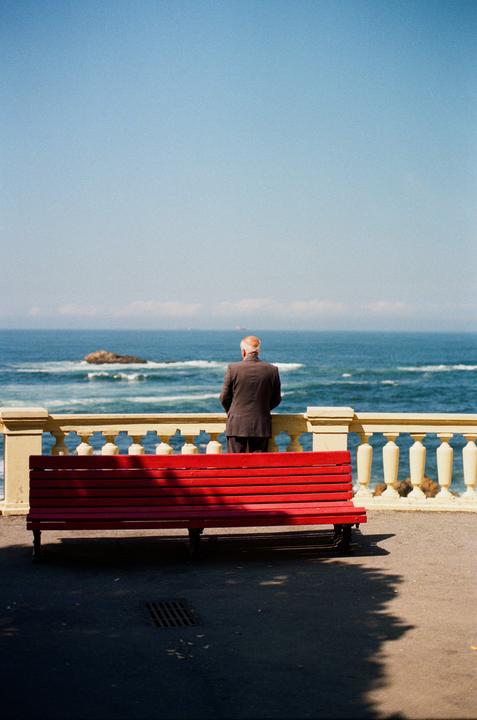 A man in a suit stands with his back to the camera, looking out at the ocean from behind a red bench and a stone railing.