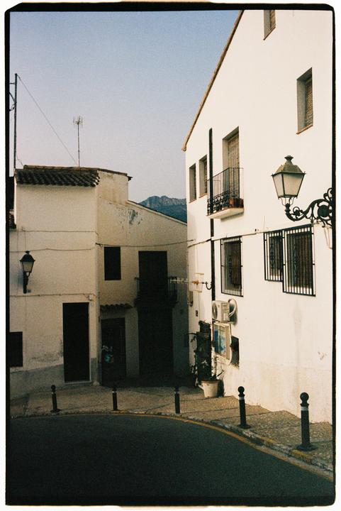 A narrow street curves past white buildings with dark windows and doors, with a mountain visible in the distance.