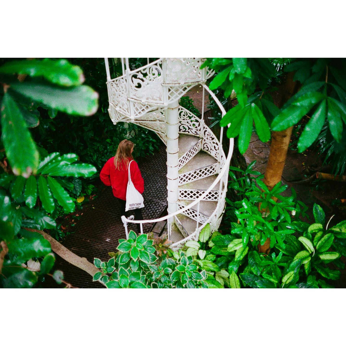 A woman in a red jacket stands next to a white spiral staircase surrounded by lush green foliage.