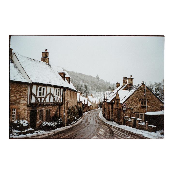 A snow-covered village street winds between stone houses with a snowy hillside in the background.